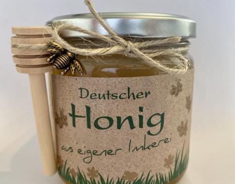 Bild von einem Glas Honig, daran hängen an einer Schleife eine kleine Biene und ein Honiglöffel