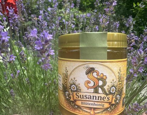 Ein Glas Blütenhonig mit der Aufschrift „Susanne’s Blütenhonig“ steht zwischen blühendem Lavendel in einem sonnigen Garten.